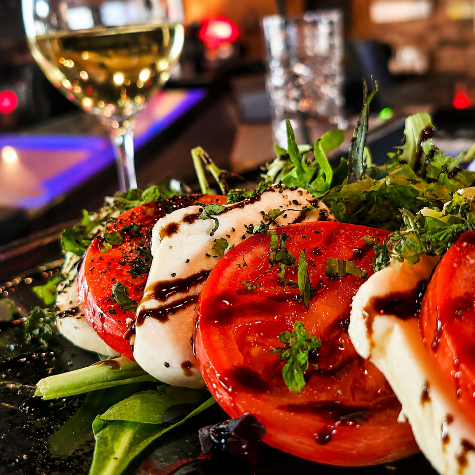 Caprese salad with ripe tomatoes, fresh mozzarella, greens, herbs, and balsamic glaze beside a glass of white wine.