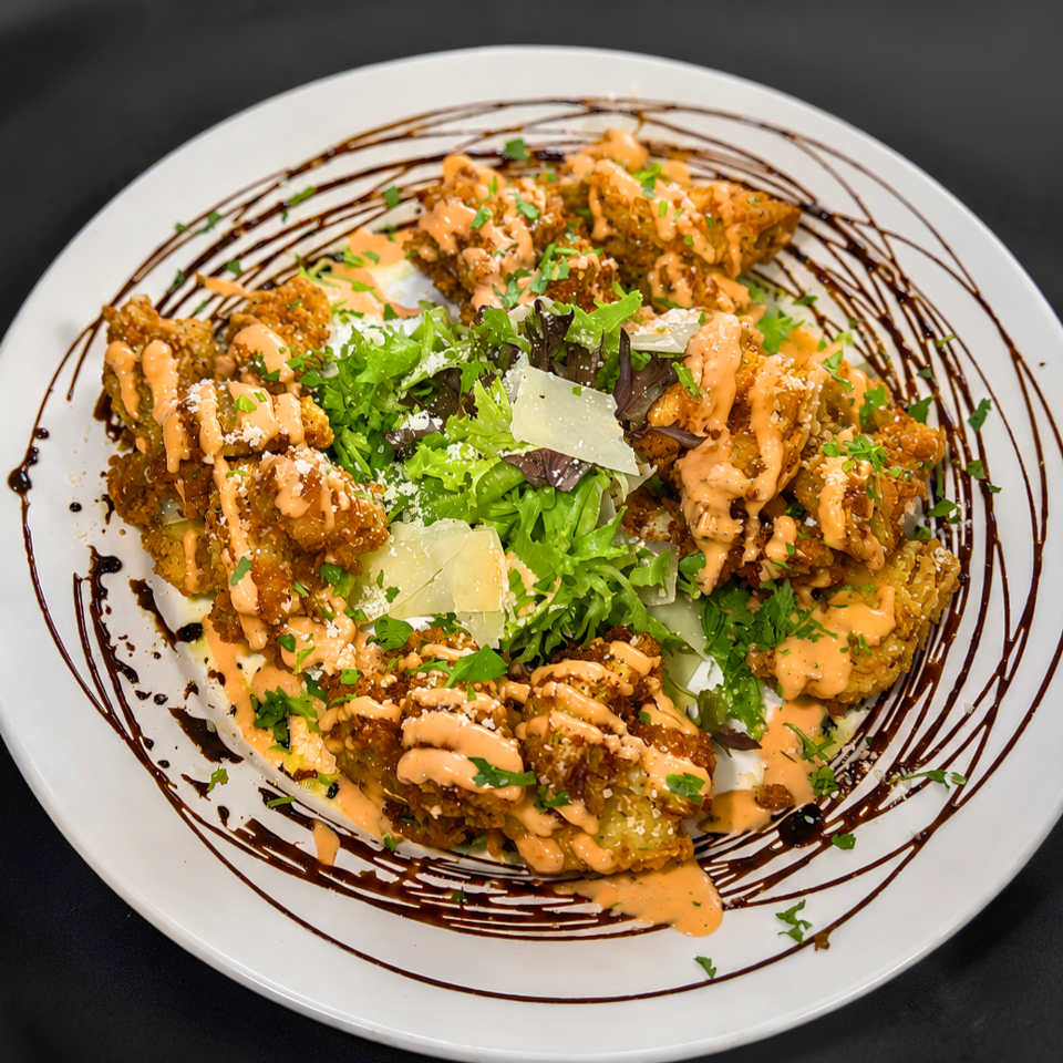 Crispy fried chicken pieces drizzled with creamy orange sauce, herbs, cheese, greens, and balsamic glaze on a white plate.
