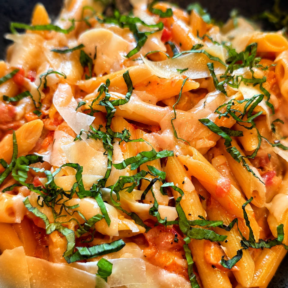 Close-up of penne pasta in creamy tomato sauce, topped with fresh basil and shaved Parmesan cheese.