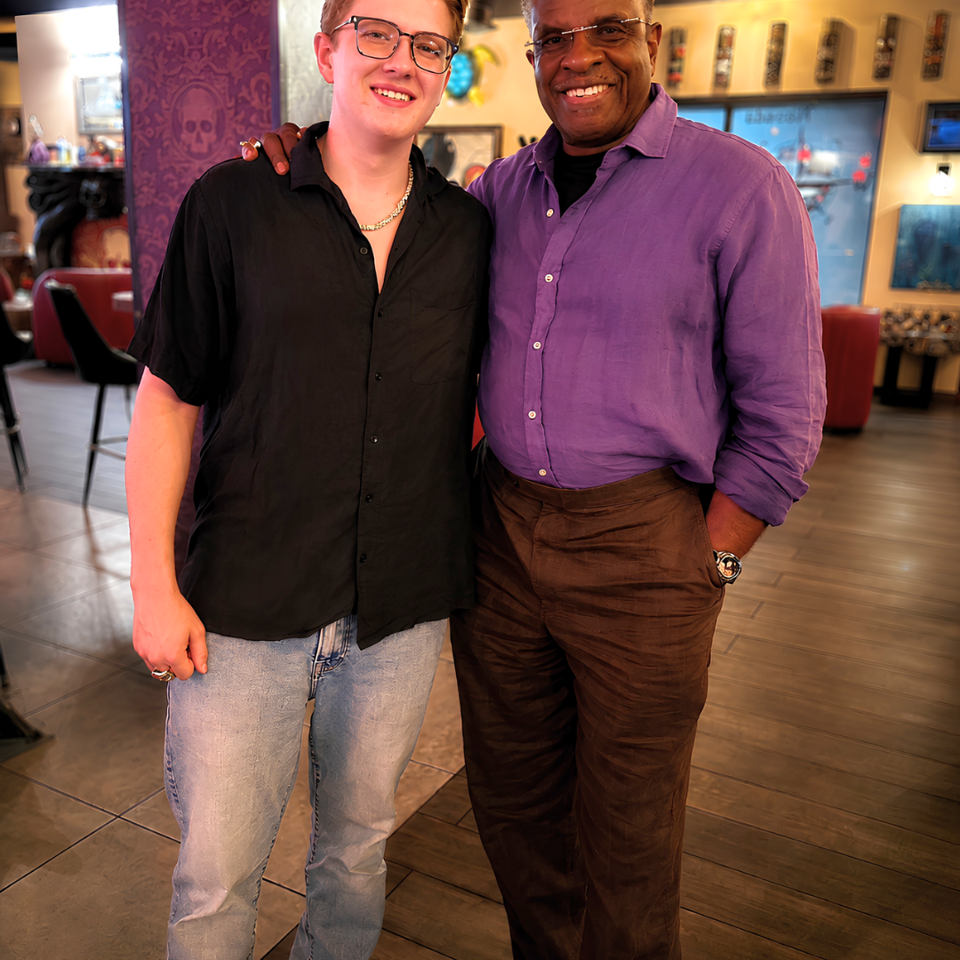 Two smiling men pose together inside a warmly lit restaurant, surrounded by colorful walls, seating, framed artwork, and televisions.