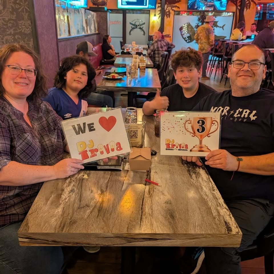 Four people pose at a restaurant table holding “We Love DJ Trivia” signs during a lively trivia night.