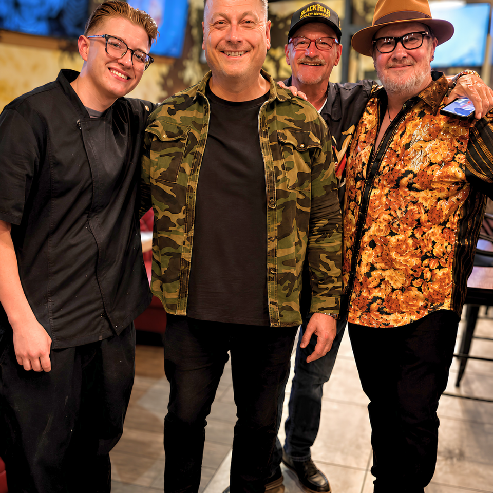Four smiling men, including a restaurant worker, pose together inside a warmly lit restaurant.