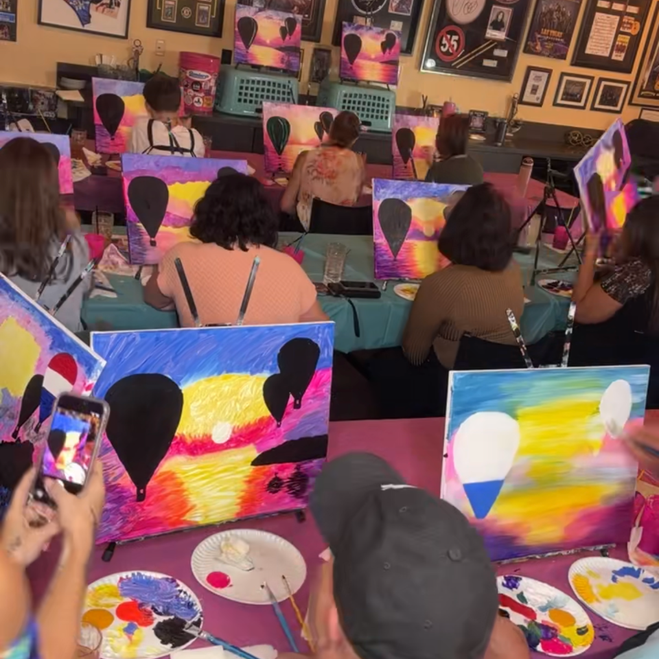 Guests paint colorful balloon-themed canvases at tables inside a lively restaurant decorated with framed sports memorabilia.