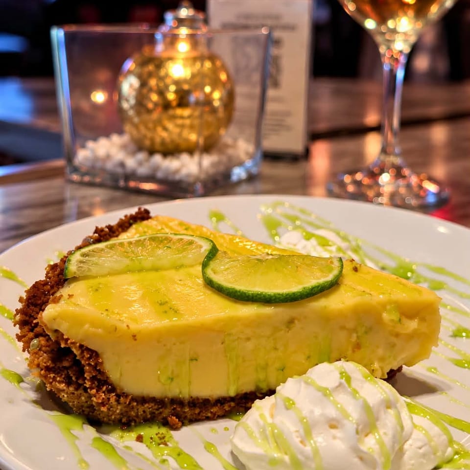 Slice of lime cheesecake with lime slices, whipped cream, and green drizzle on a plate in a warmly lit restaurant.