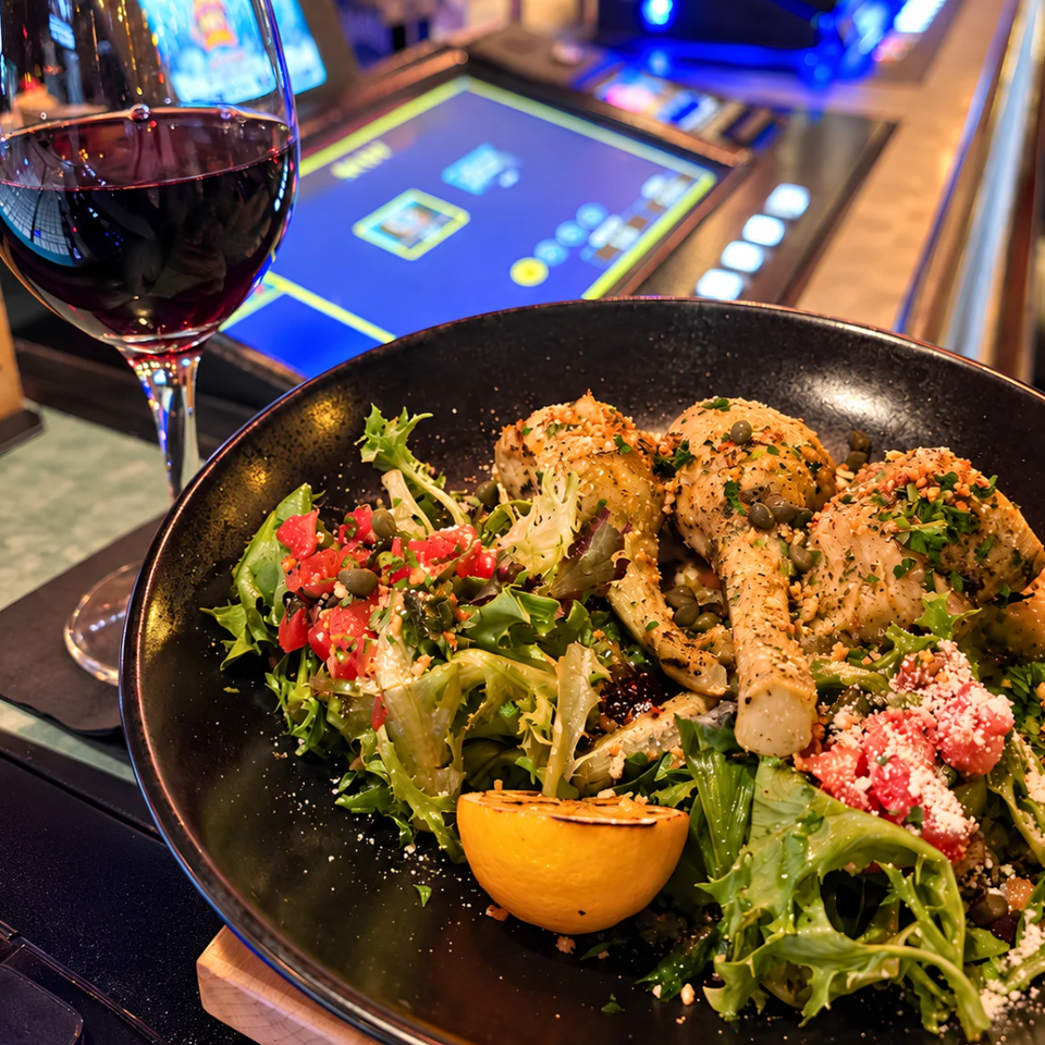 Gourmet salad with grilled chicken, greens, tomatoes, feta, and lemon beside a glass of red wine at a restaurant bar.