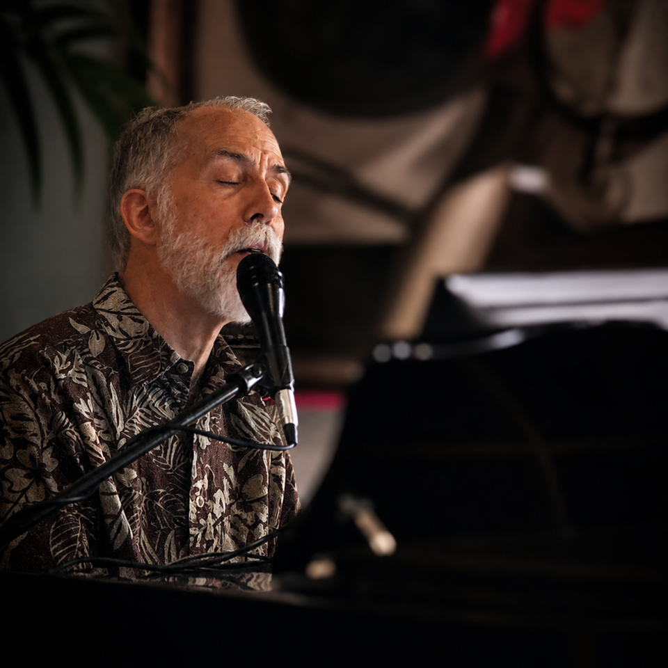 Gray-haired musician singing into a microphone while playing piano in a dimly lit restaurant.