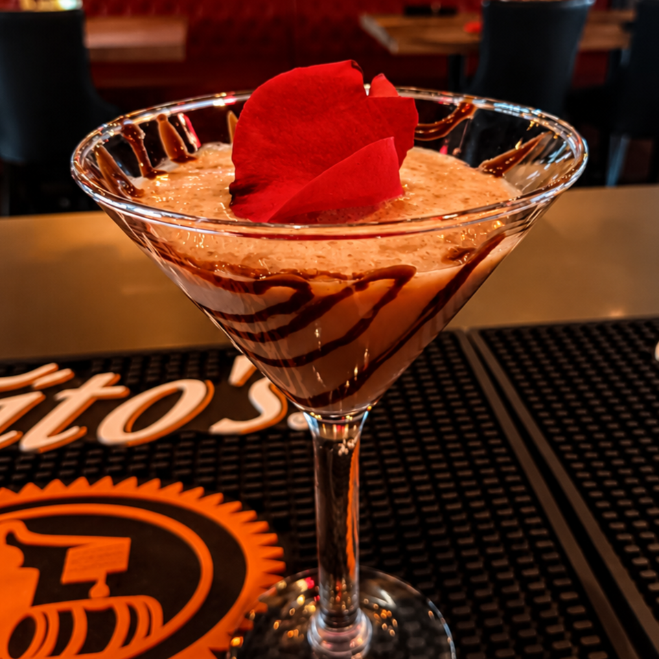 Chocolate-drizzled dessert cocktail in a martini glass, topped with a red rose petal, sits on a restaurant bar.