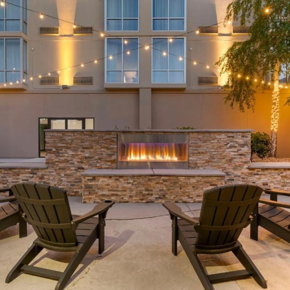 Outdoor courtyard with Adirondack chairs surrounding a stone fire feature, illuminated by string lights and a nearby tree.