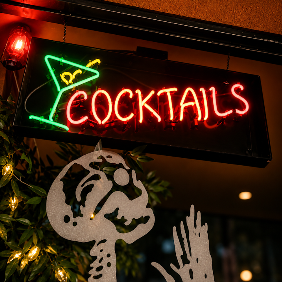 Glowing neon cocktail sign hangs above leafy indoor décor and a metal skeleton ornament in a dimly lit bar.