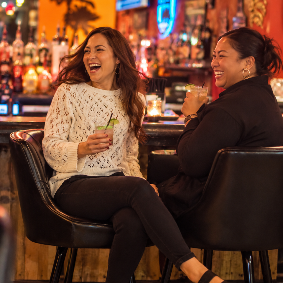 Two women laugh together in a warmly lit bar, each holding a cocktail amid colorful bottles and neon décor.