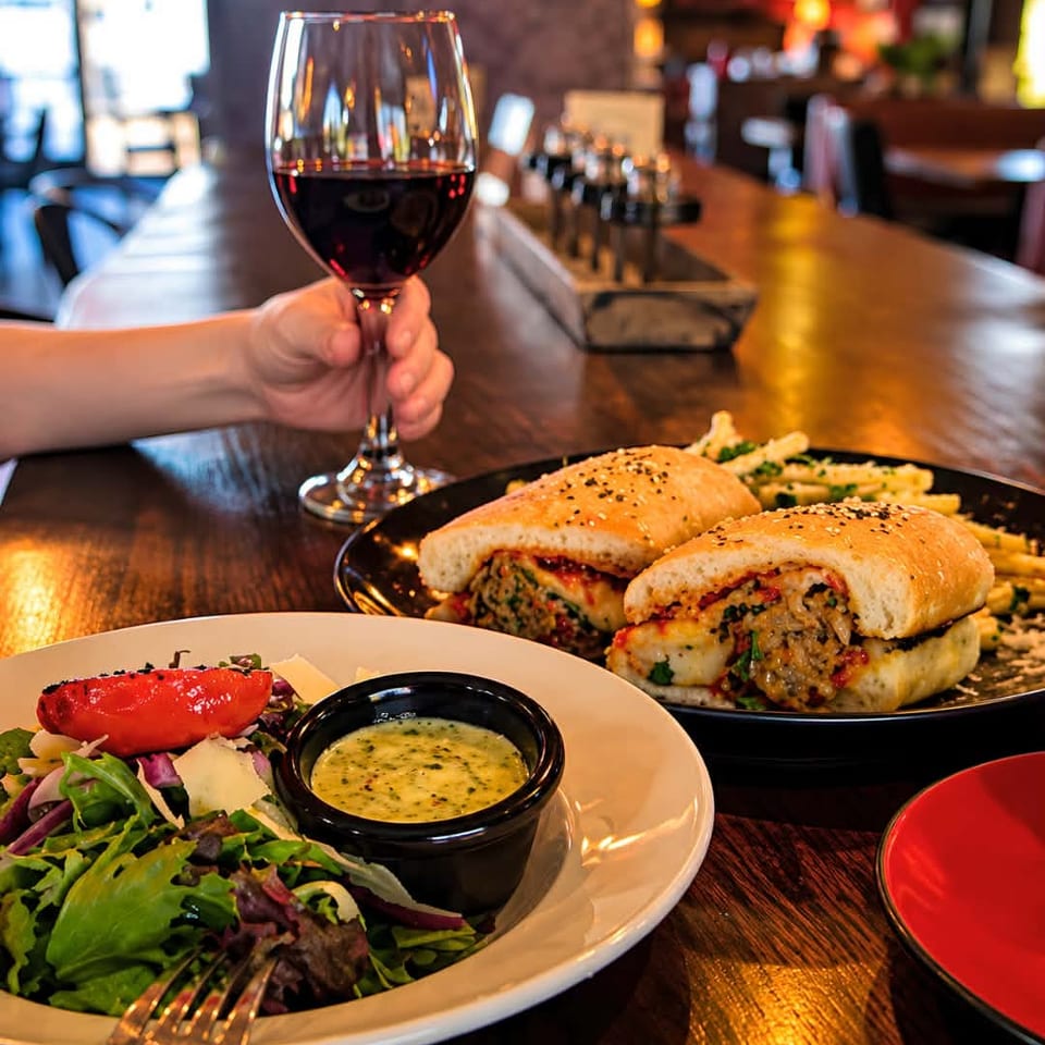 Restaurant table featuring a glass of red wine, toasted sandwiches with fries, and a salad with dipping sauce.