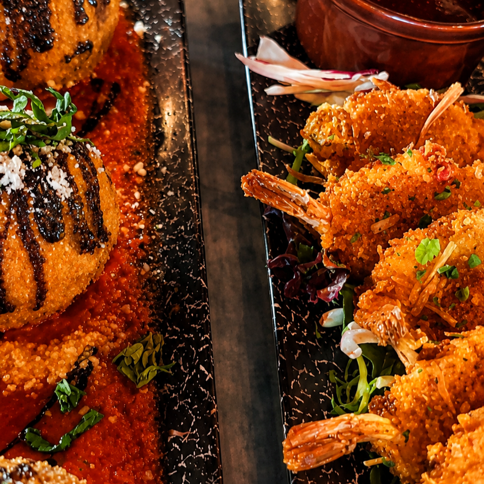 Two black plates feature crispy breaded shrimp, sauced fried appetizers, garnishes, and a cup of dipping sauce.