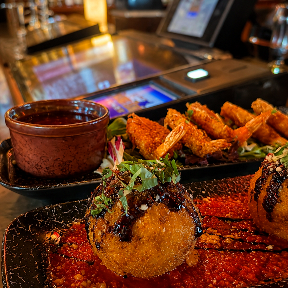 Golden fried shrimp and croquettes served with red sauce, greens, and dipping sauce at a warmly lit restaurant bar.