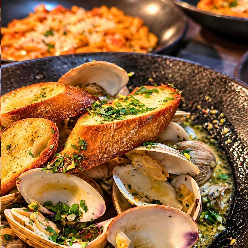 Garlic bread and open clams garnished with herbs in a skillet, with pasta dishes visible in the background.