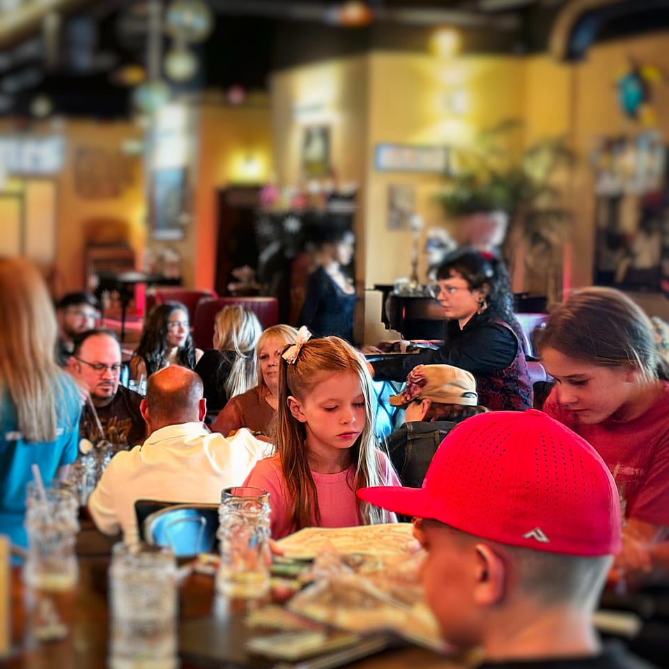 Busy restaurant dining room filled with families and groups seated at tables, with menus, drinks, and warm ambient lighting.