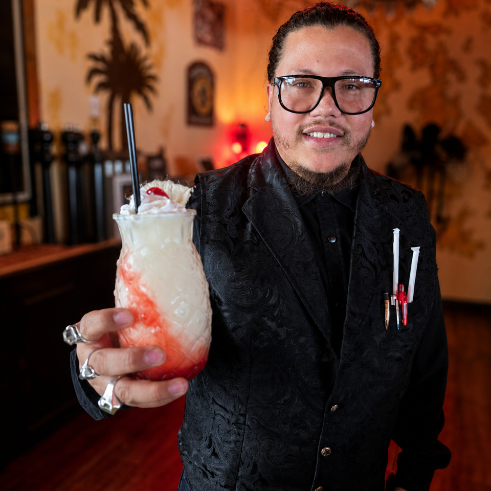 Smiling bartender in a black patterned suit presents a whipped cream-topped strawberry cocktail inside a warmly lit restaurant.